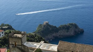 A coastal view with a castle on a cliff, a boat creating a wake, and buildings with tiled roofs. - VivimiHome-Casa Tiberio (Conca dei Marini)