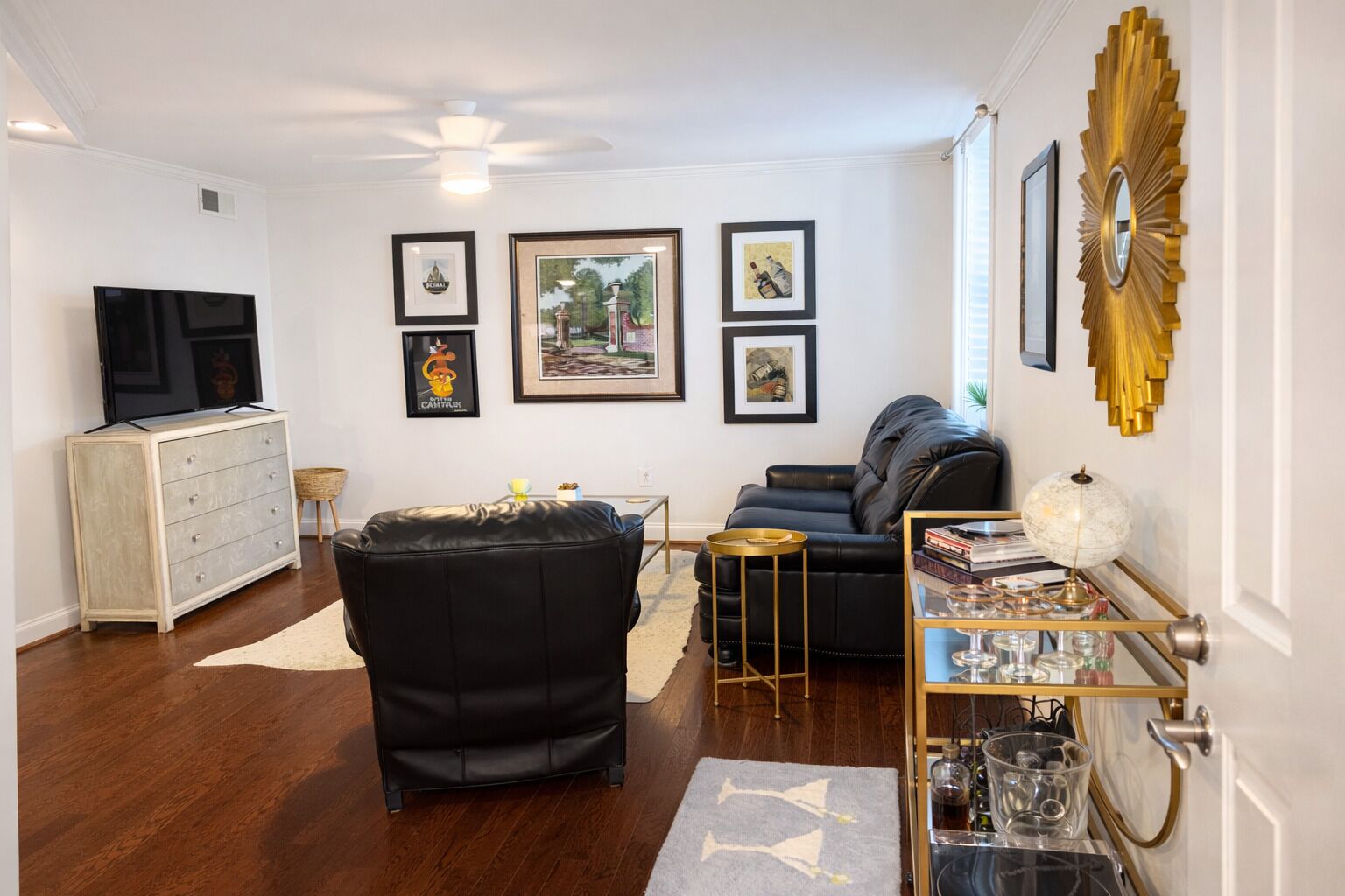 Un salon avec un canapé en cuir noir, un meuble blanc, un téléviseur, des tableaux encadrés au mur et un miroir décoratif.