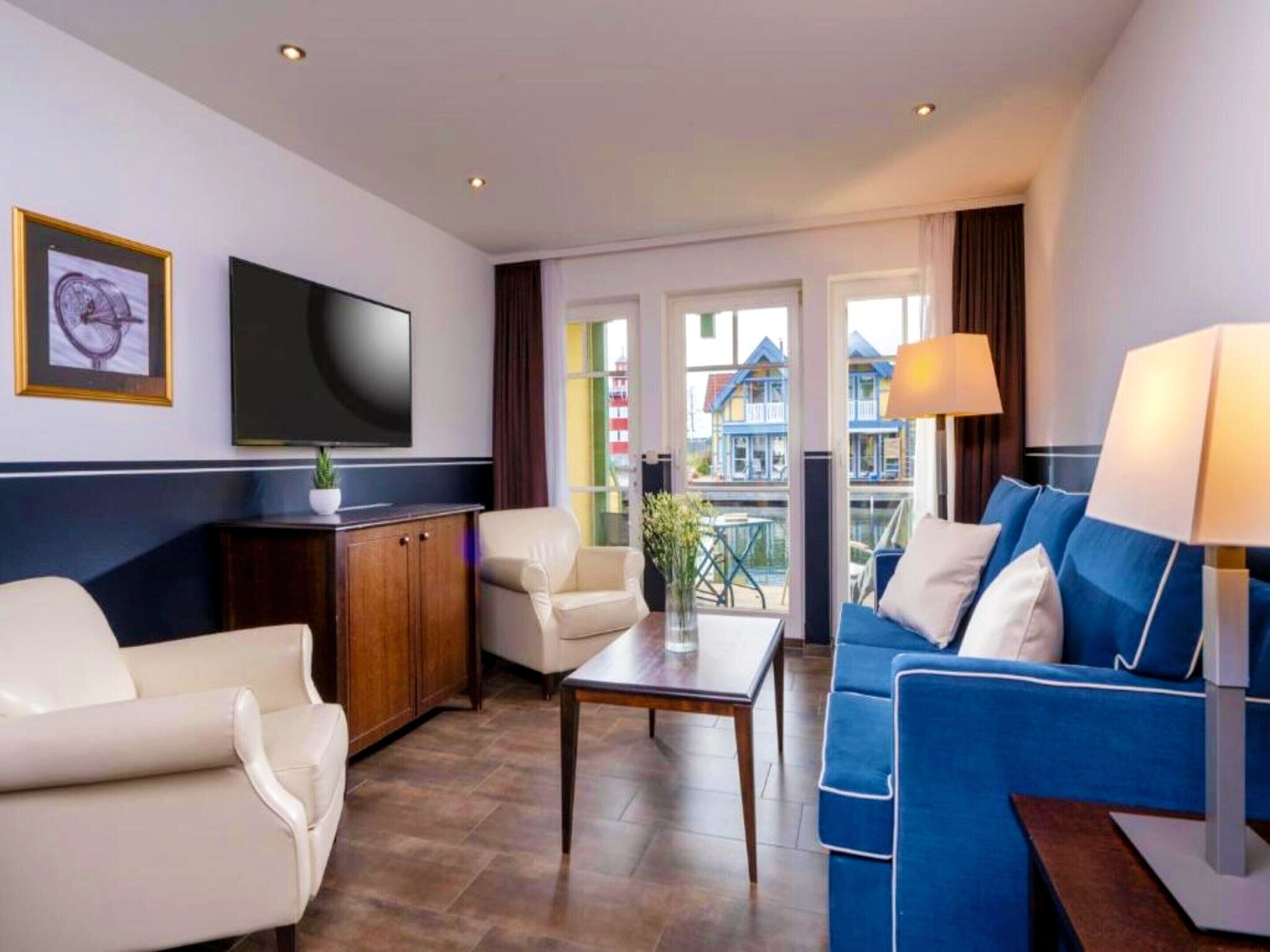 Habitación de hotel con televisor, sofá, sillón, mesa y vistas a edificios a través de las ventanas.