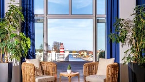 Ein Zimmer mit zwei Korbstühlen, einem kleinen runden Tisch und großen Fenstern mit Blick auf einen Leuchtturm und Gebäude über dem Wasser.