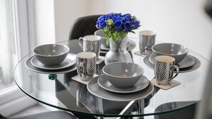 Una mesa de comedor con cubierta de vidrio, rodeada de cuencos grises y tazas con diseños, y un florero con flores azules.