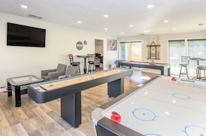 Ein Billardtisch, ein Kicker und ein Shuffleboard-Tisch in einem gut beleuchteten Raum mit einem Fernseher an der Wand.