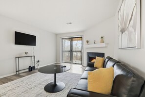 Ein modernes Wohnzimmer mit einer schwarzen Ledersofa, einem runden Couchtisch, einem flachbildfernseher an der Wand, einem Kamin und einem großen Fenster mit Jalousien.