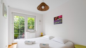Un dormitorio con una cama grande, dos almohadas, una mesita de noche con una lámpara, una ventana con vista a la vegetación y un cuadro en la pared.