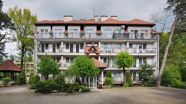 Un edificio de varios pisos con balcones, un vestíbulo cubierto y un jardín con árboles y arbustos.
