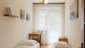 Habitación de hotel con dos camas individuales, suelo de madera, ventana con cortinas translúcidas, una mesita y un cuadro en la pared.