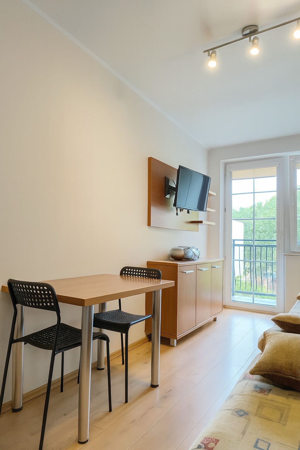 Uma sala de estar moderna com uma mesa de jantar de madeira, duas cadeiras pretas, um sofá e uma televisão fixada na parede.