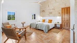 Een ruime kamer met een groot bed, twee nachtkastjes, een stoel, een klein tafeltje met bloemen, en uitzicht op groen buiten.