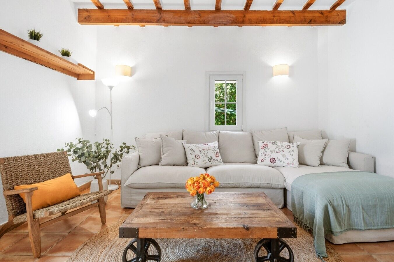 Ein Wohnzimmer mit einer Couch, einem Holztisch, einem Korbstuhl und einem Fenster mit Blick auf Grünflächen.