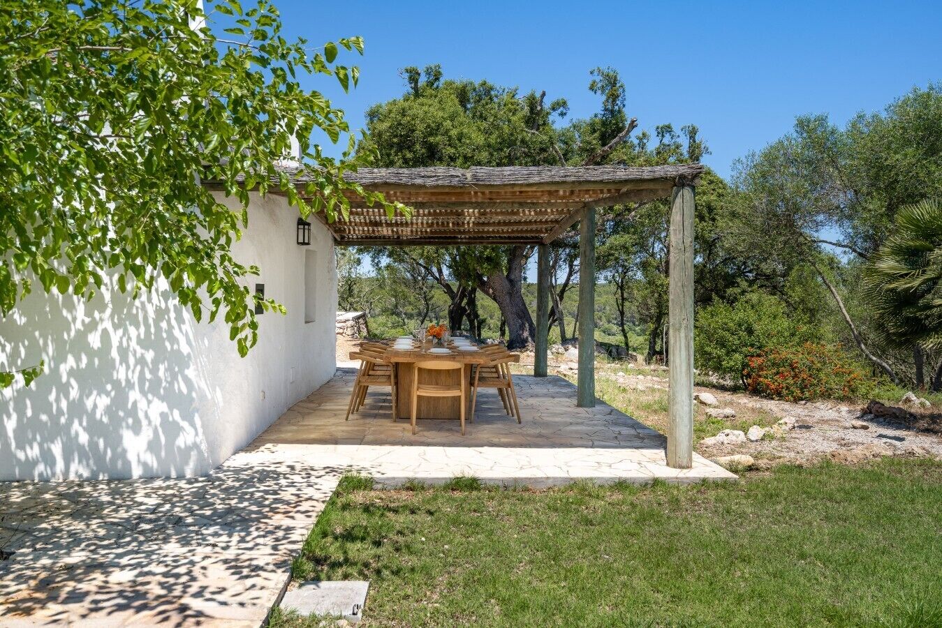 Un espace extérieur abrité, aménagé pour les repas, avec une table et des chaises, entouré d’un bâtiment blanc et d’un paysage naturel.
