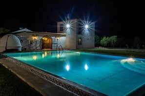 Una piscina con fachada de piedra, zona cubierta e iluminación exterior durante la noche.