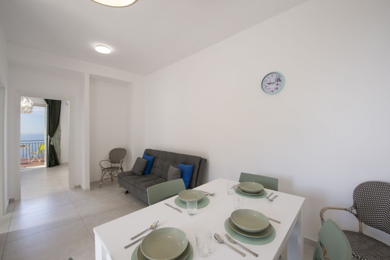 A dining area with a table set for four, a grey sofa with blue cushions, and a view of the sea through an open door.