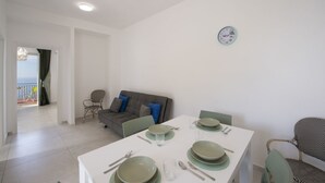 Et spiseområde med et bord dækket til fire personer, en grå sofa med blå hynder, og havudsigt gennem en åben dør.