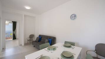 A dining area with a table set for four, a grey sofa with blue cushions, and a view of the sea through an open door.