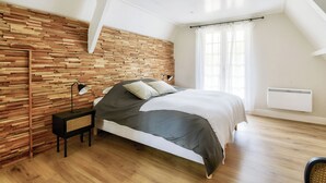 Quarto com cabeceira de madeira, cama com roupa de cama cinza, mesa de cabeceira com abajur e janela com cortinas.