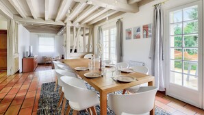Uma sala de jantar com uma mesa de madeira posta para seis, cadeiras brancas e uma janela grande com cortinas.