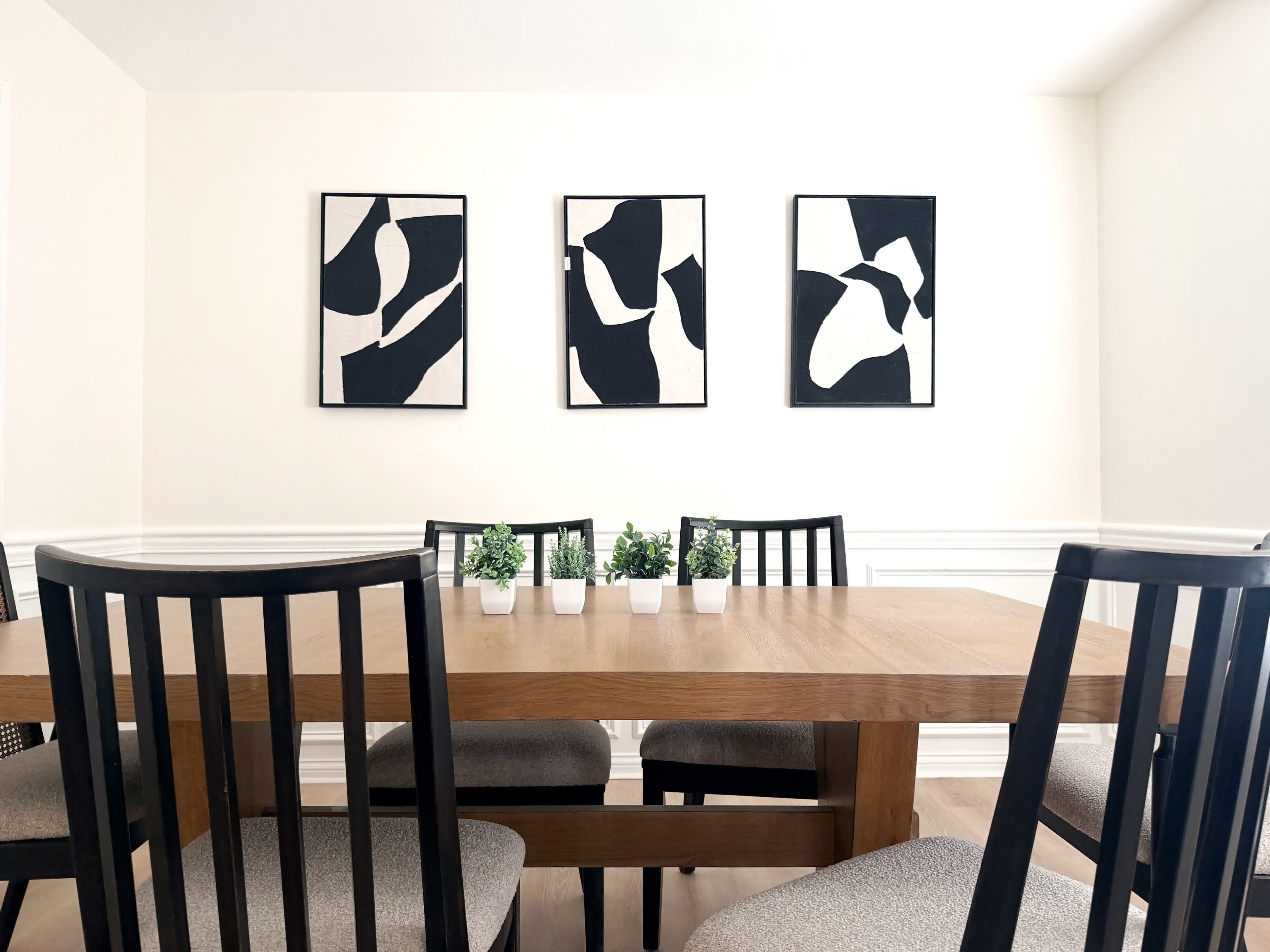 Een eetruimte met een houten tafel, vier stoelen en drie abstracte zwart-wit schilderijen aan de muur.
