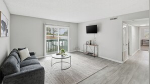 Un salon moderne avec un canapé gris, une table basse ronde agrémentée d’une plante, un téléviseur fixé au mur et une porte-fenêtre coulissante donnant sur un balcon.
