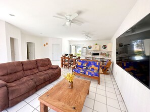 Sala de estar com um sofá marrom, uma mesa de centro de madeira com um vaso de flores, um sofá azul estampado e uma área de cozinha com armários e uma geladeira.