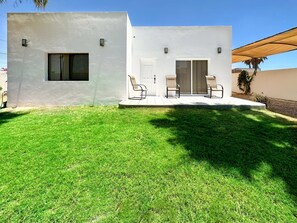 Uma casa moderna de dois andares com fachada branca, varanda coberta com duas cadeiras e gramado bem cuidado.