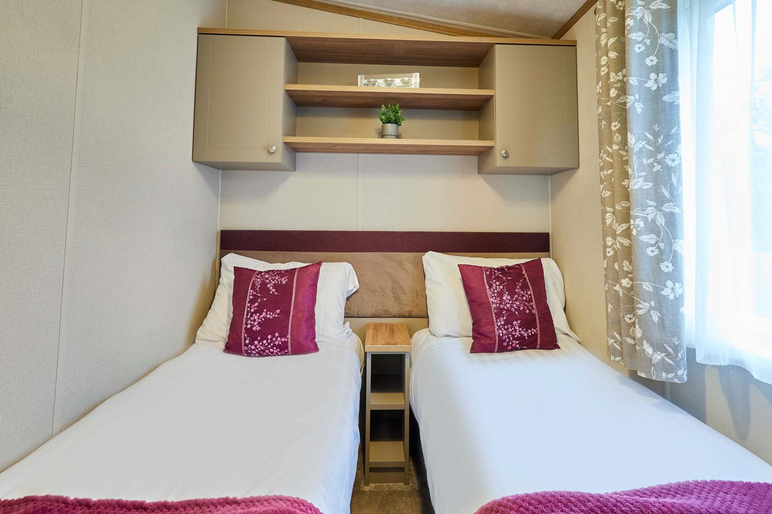 A room with two beds, a small shelf above the beds, and a window with floral curtains.