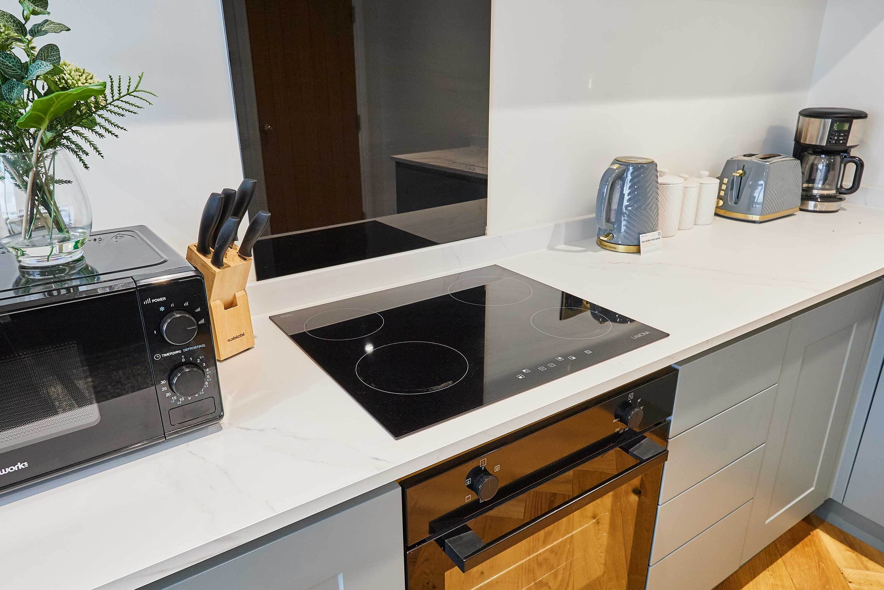 Una cocina con microondas negro, placa de inducción integrada, bloque de cuchillos, cafetera y tostadora.