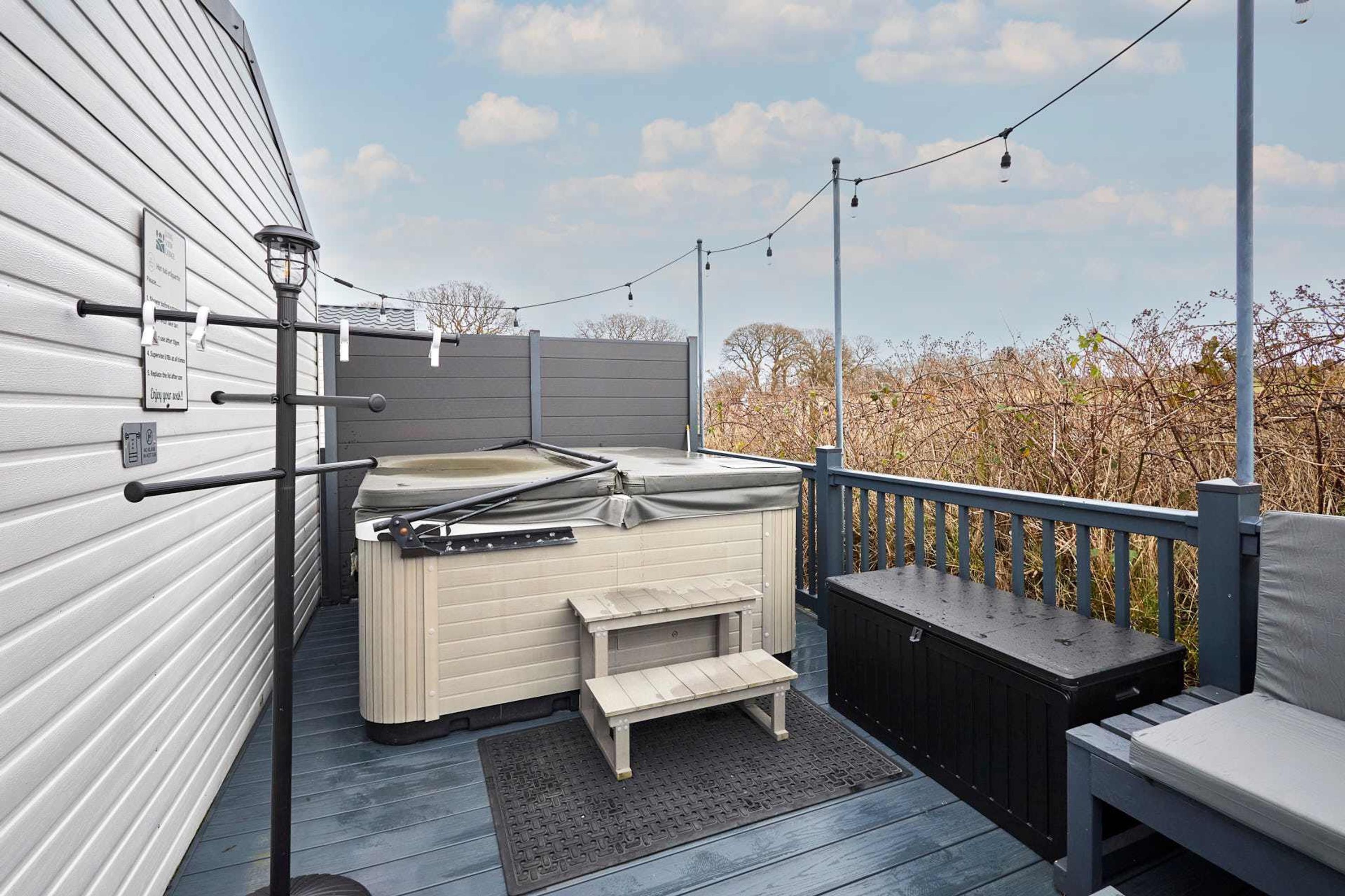 A hot tub on a deck with a wooden bench and a small table, surrounded by a railing and string lights.