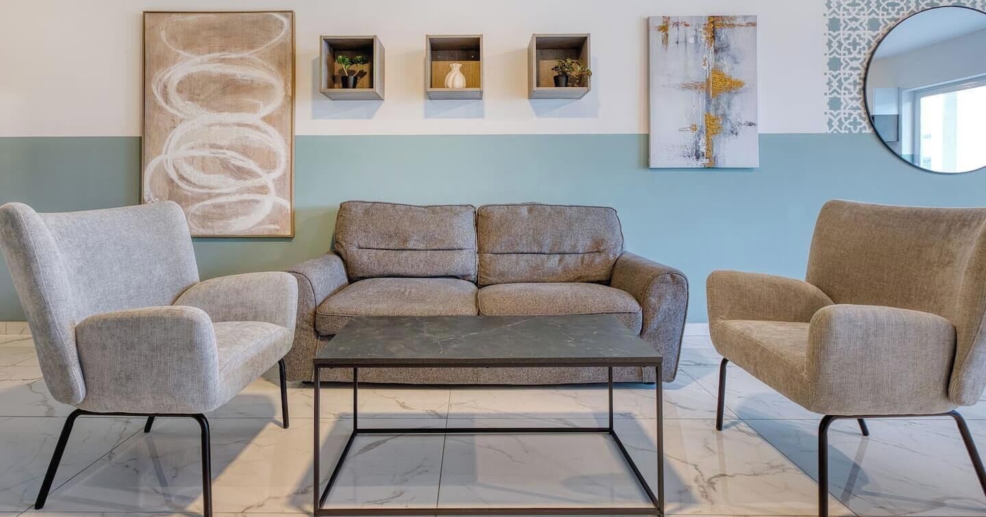A modern living room with a grey sofa, two armchairs, and a coffee table. There are three wall-mounted shelves with decorative items, a large abstract painting, and a round mirror.