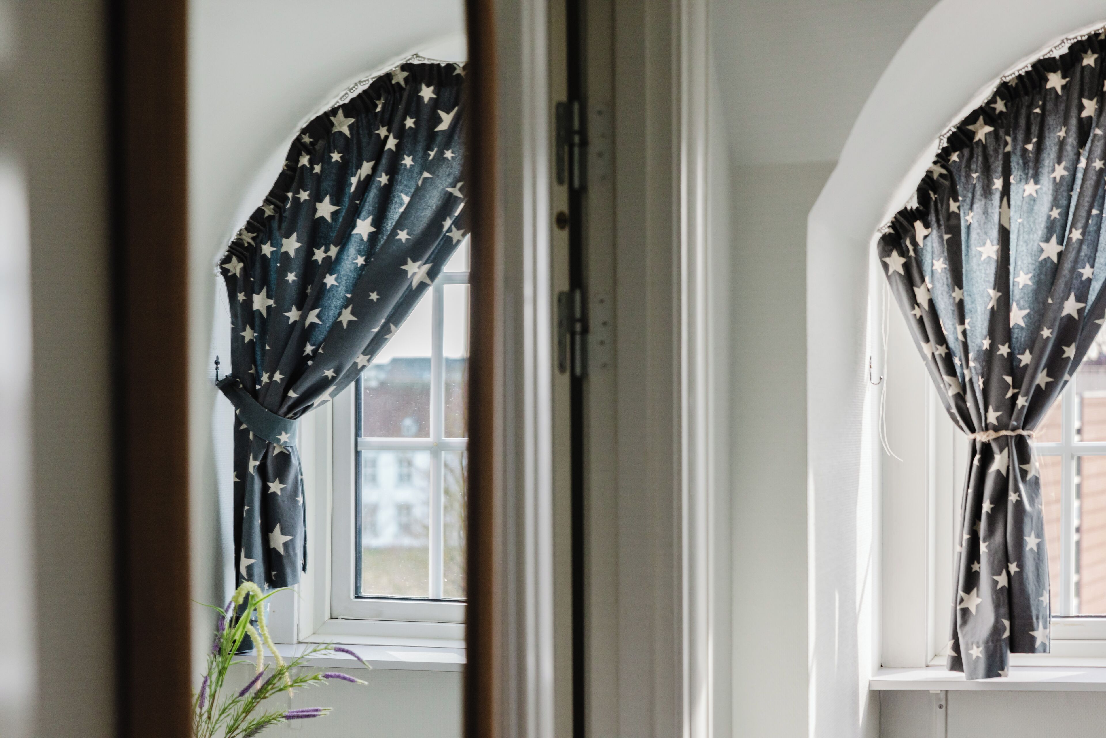 Una habitación con ventana, cortinas con estampado de estrellas, una puerta blanca y vista a un edificio exterior.