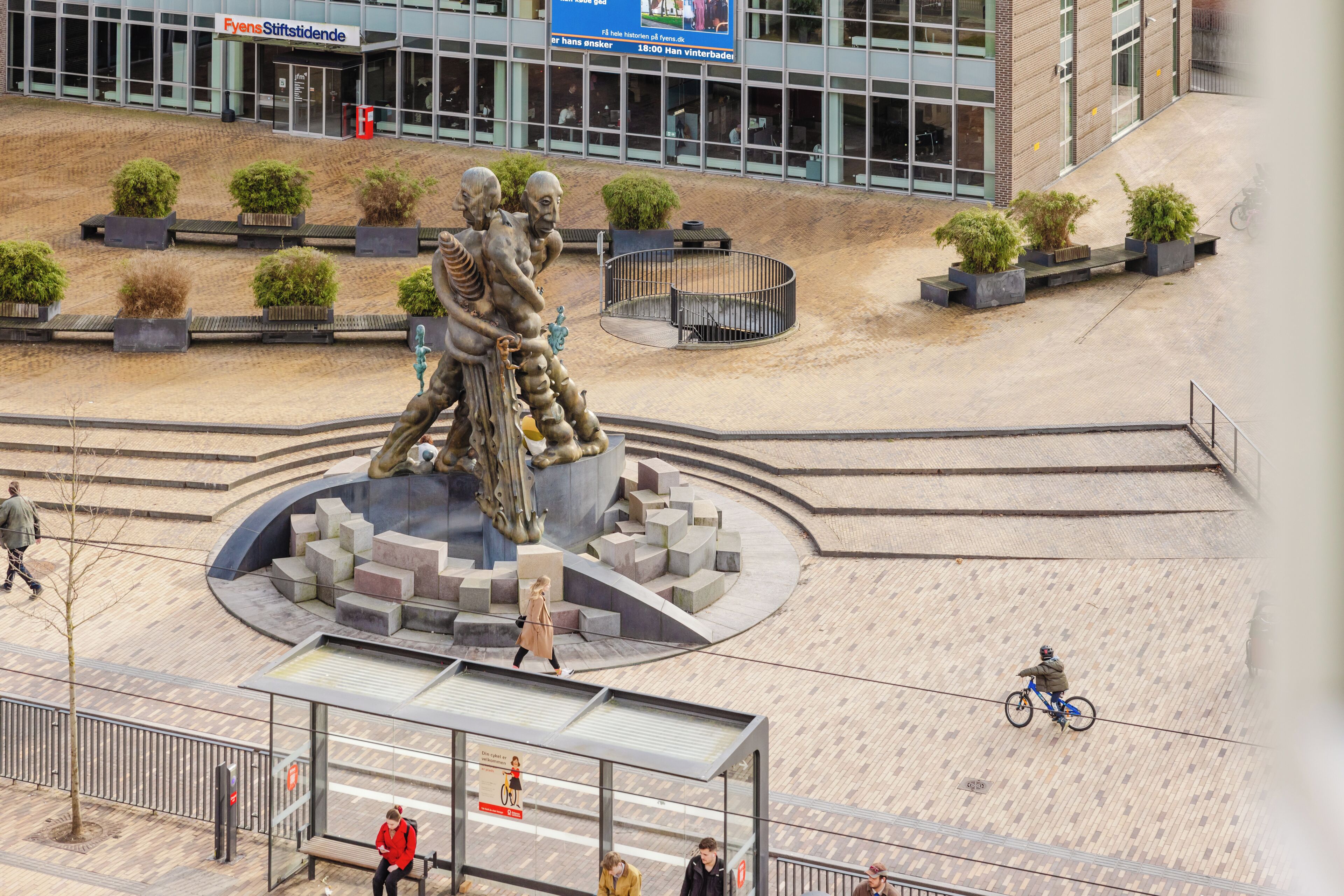A public square with a central sculpture, benches, and a building in the background.