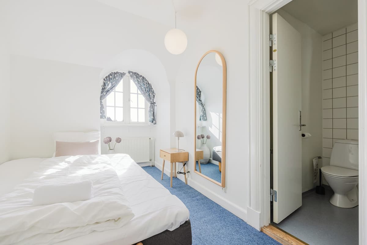 A bedroom with a bed, a bedside table, a mirror, a window with curtains, and a bathroom with a toilet and sink.