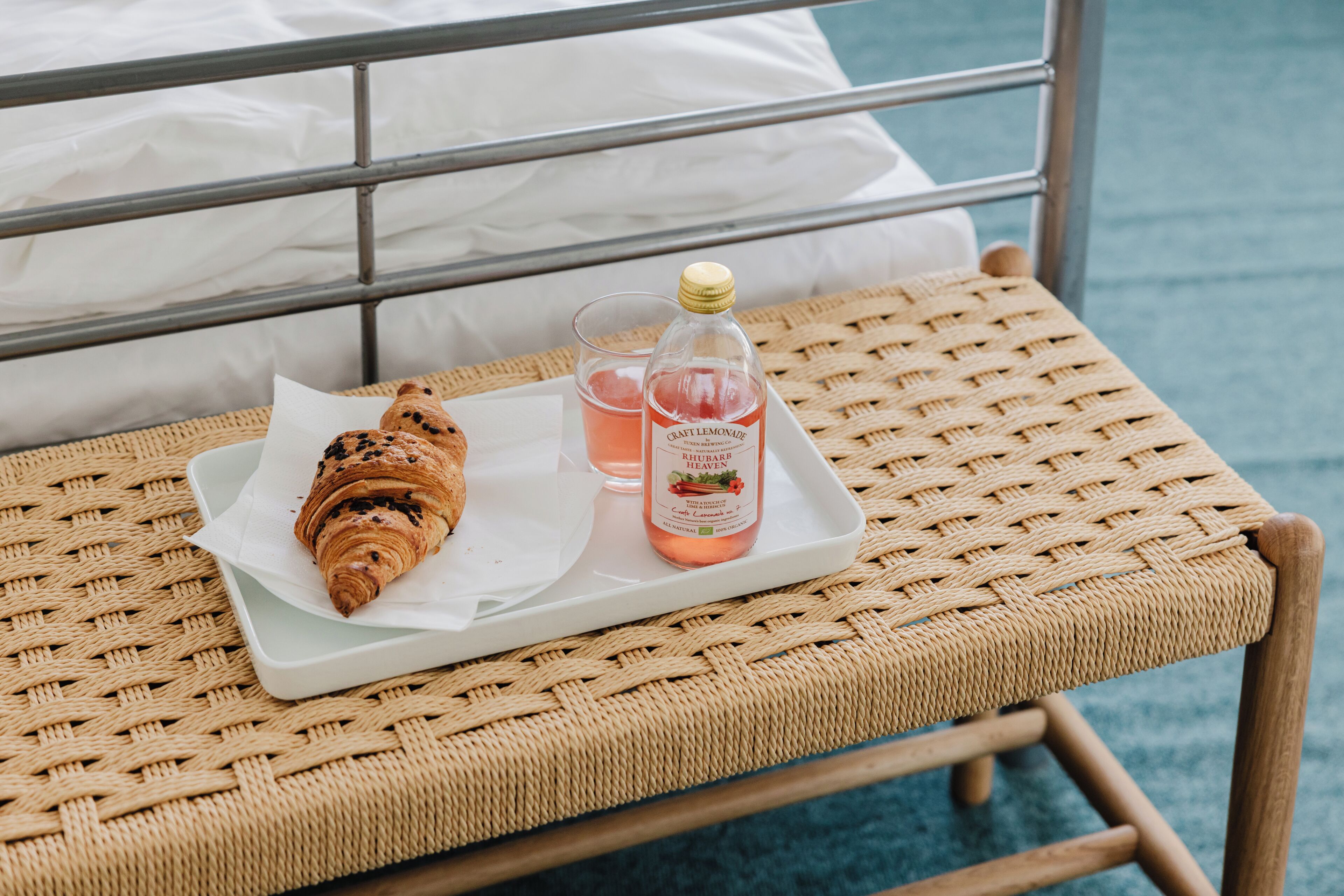 Una poltrona in vimini con un vassoio contenente un cornetto, un bicchiere di bevanda rosa e una bottiglia di limonata.