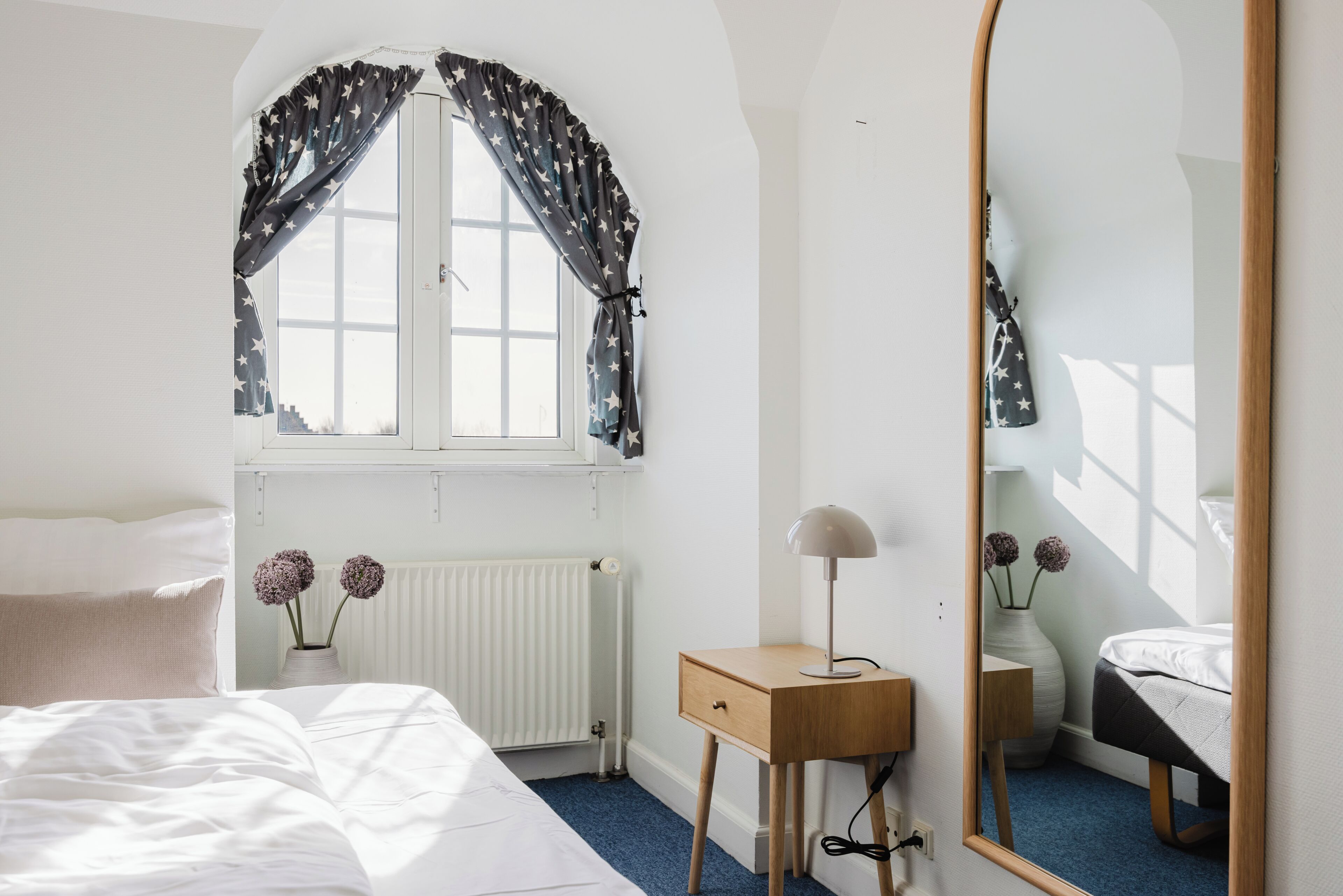 A bedroom with a bed, a bedside table with a lamp, a mirror, a window with star-patterned curtains, and a radiator.