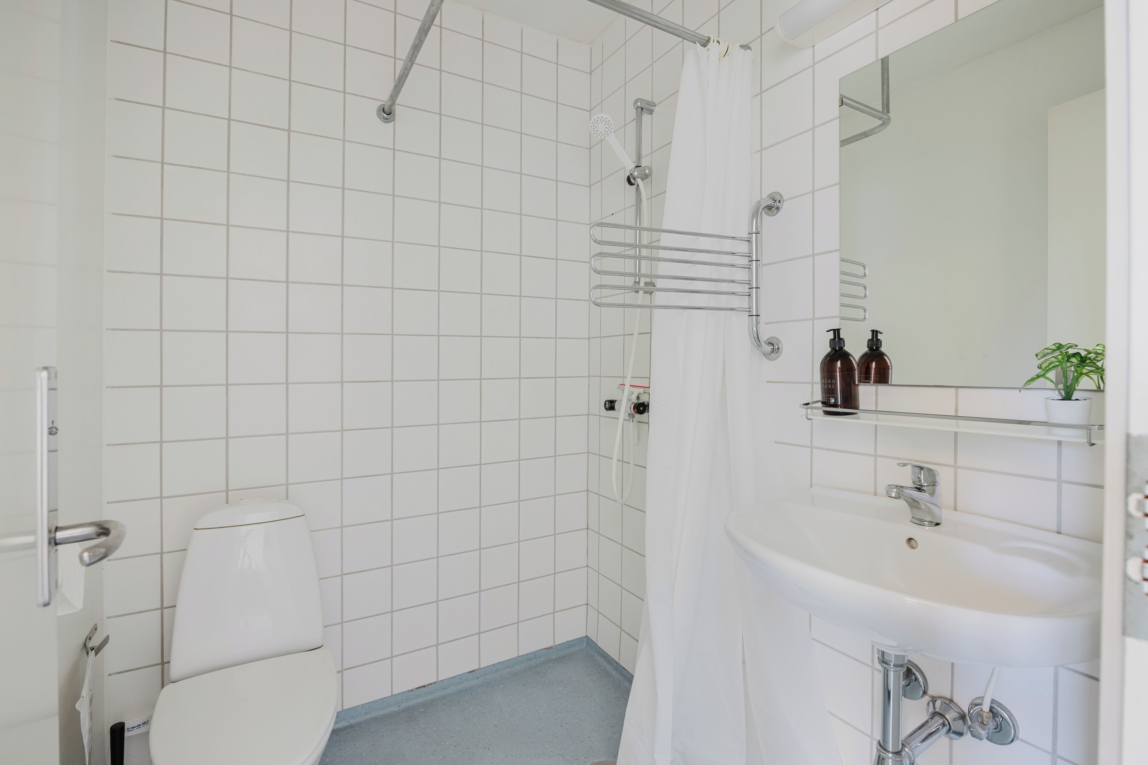 A bathroom with white tiled walls, a shower, a toilet, and a sink with a mirror above it.