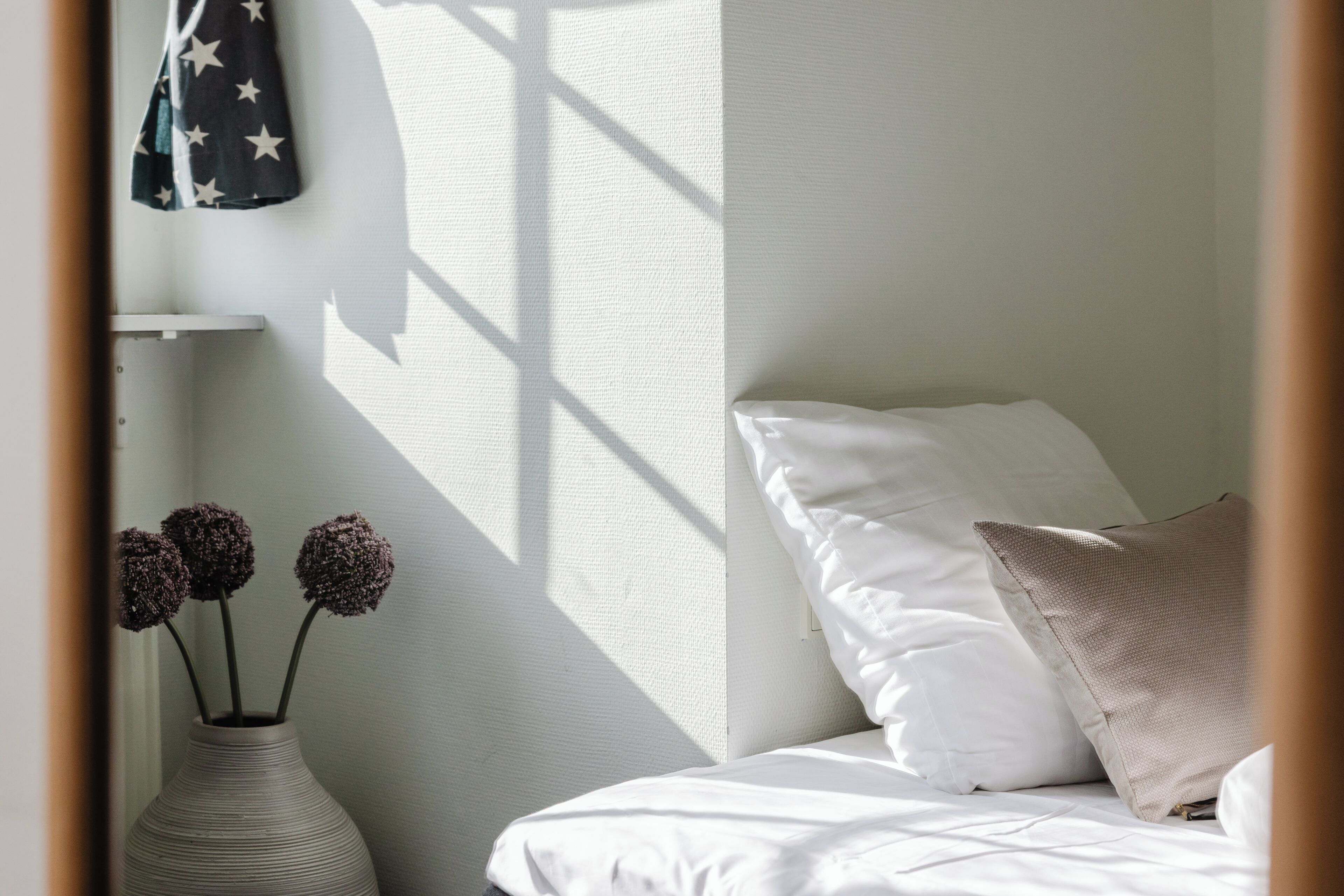 Una cama con ropa blanca, una almohada color beige y un florero blanco con flores sobre una mesita de noche.