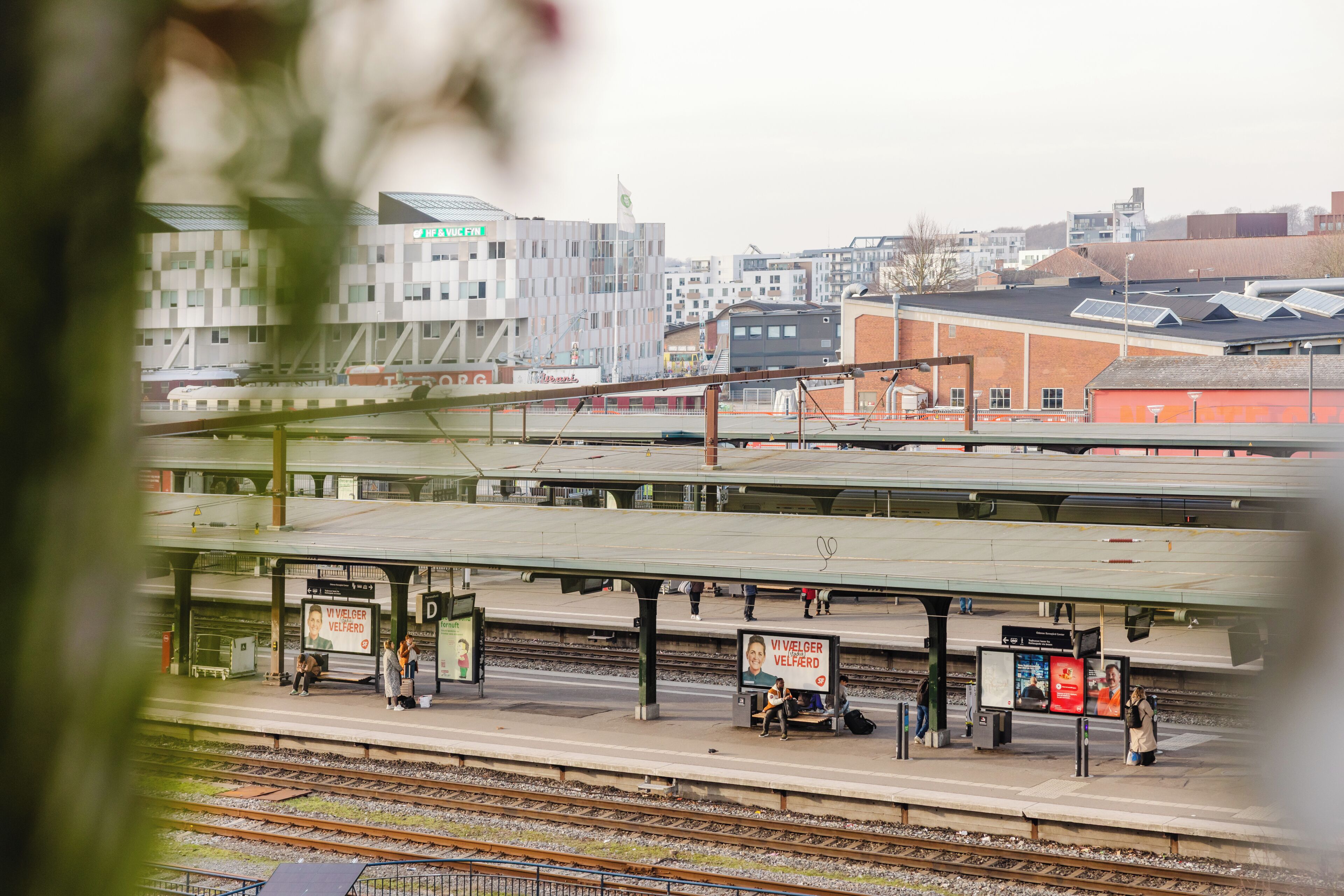 A train station platform with multiple tracks, overhead lines, and a few passengers.