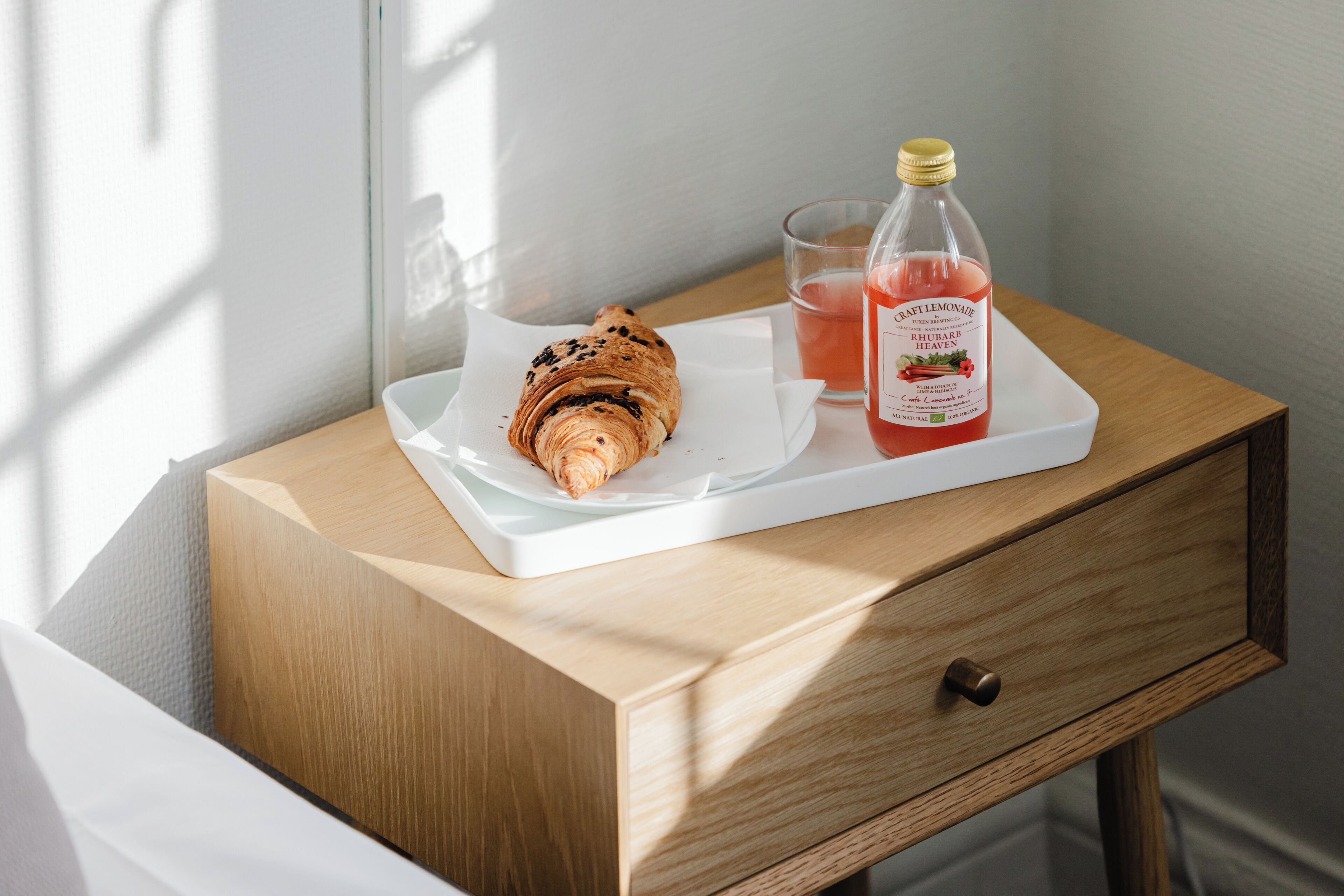 Una mesita de noche de madera con una bandeja blanca que sostiene un croissant y una botella de limonada.