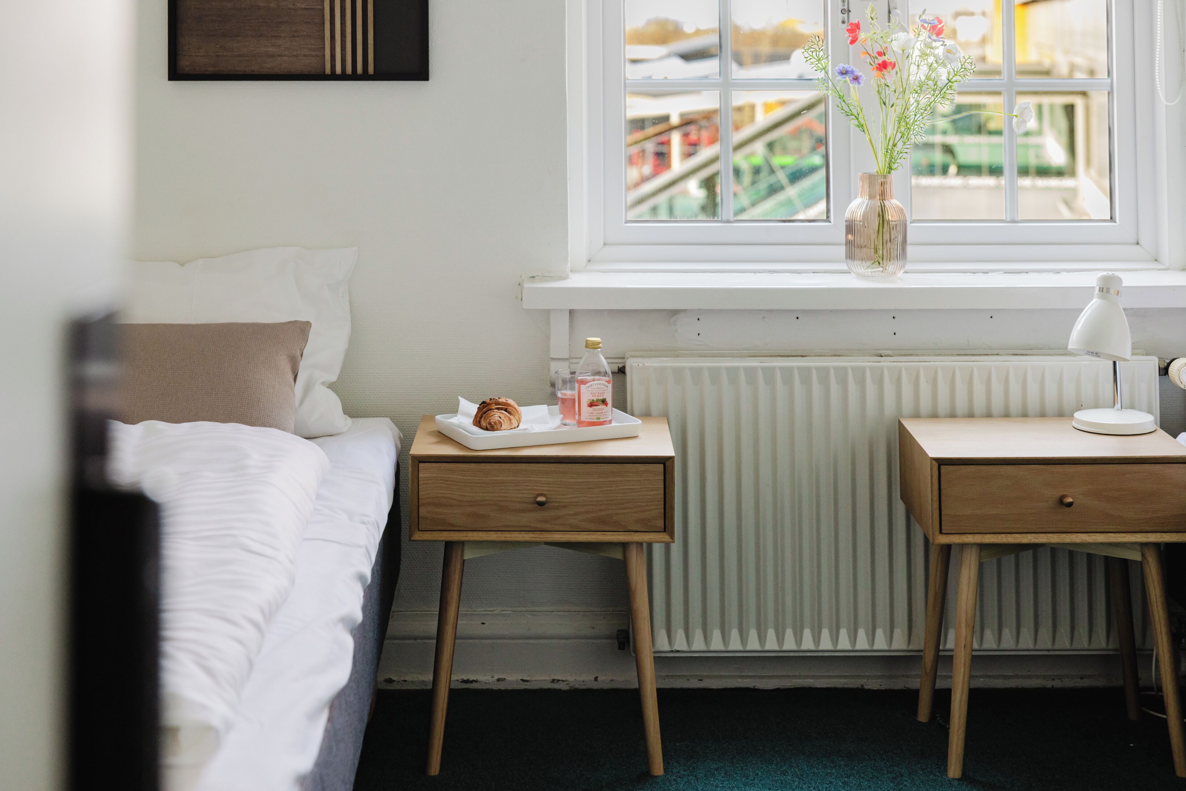 Une chambre à coucher avec un lit, une table de chevet sur laquelle se trouve un plateau de nourriture, une petite table avec une lampe, et une fenêtre donnant sur un immeuble.
