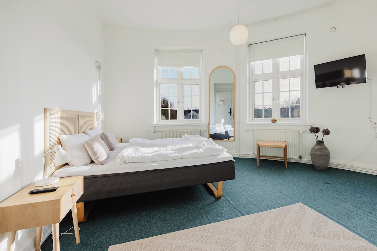 A bedroom with a bed, a desk, a chair, a TV, a window, and a mirror.