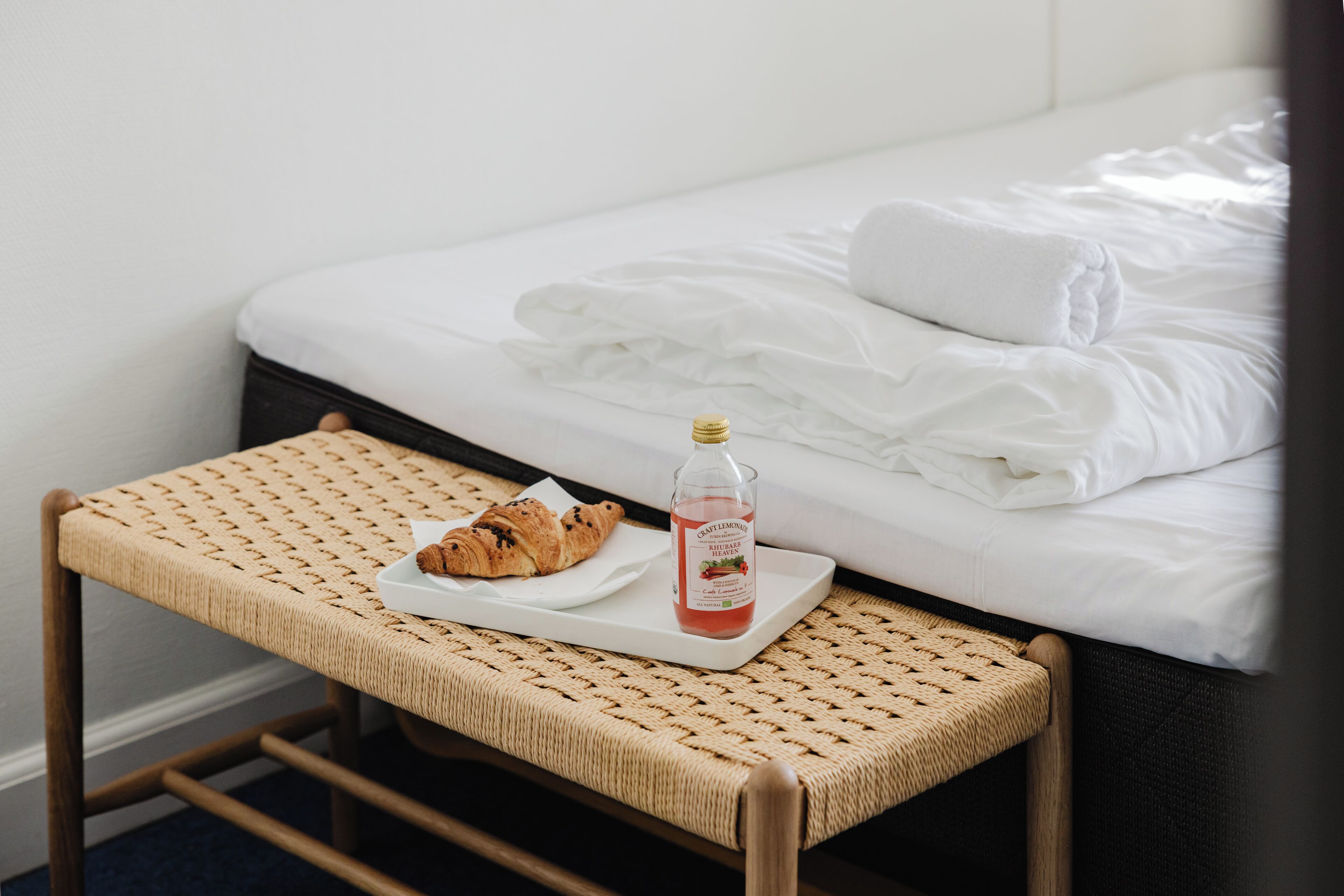 Un letto con lenzuola bianche, una sedia in vimini con un vassoio che contiene un cornetto e una bottiglia di succo, e una testiera in legno scuro.