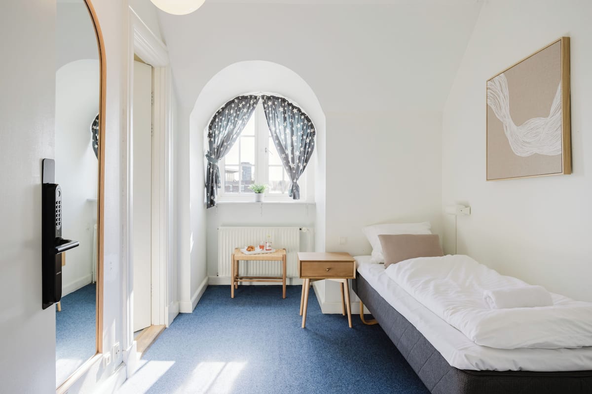 A bedroom with a bed, a desk, a chair, a window with curtains, and a radiator.