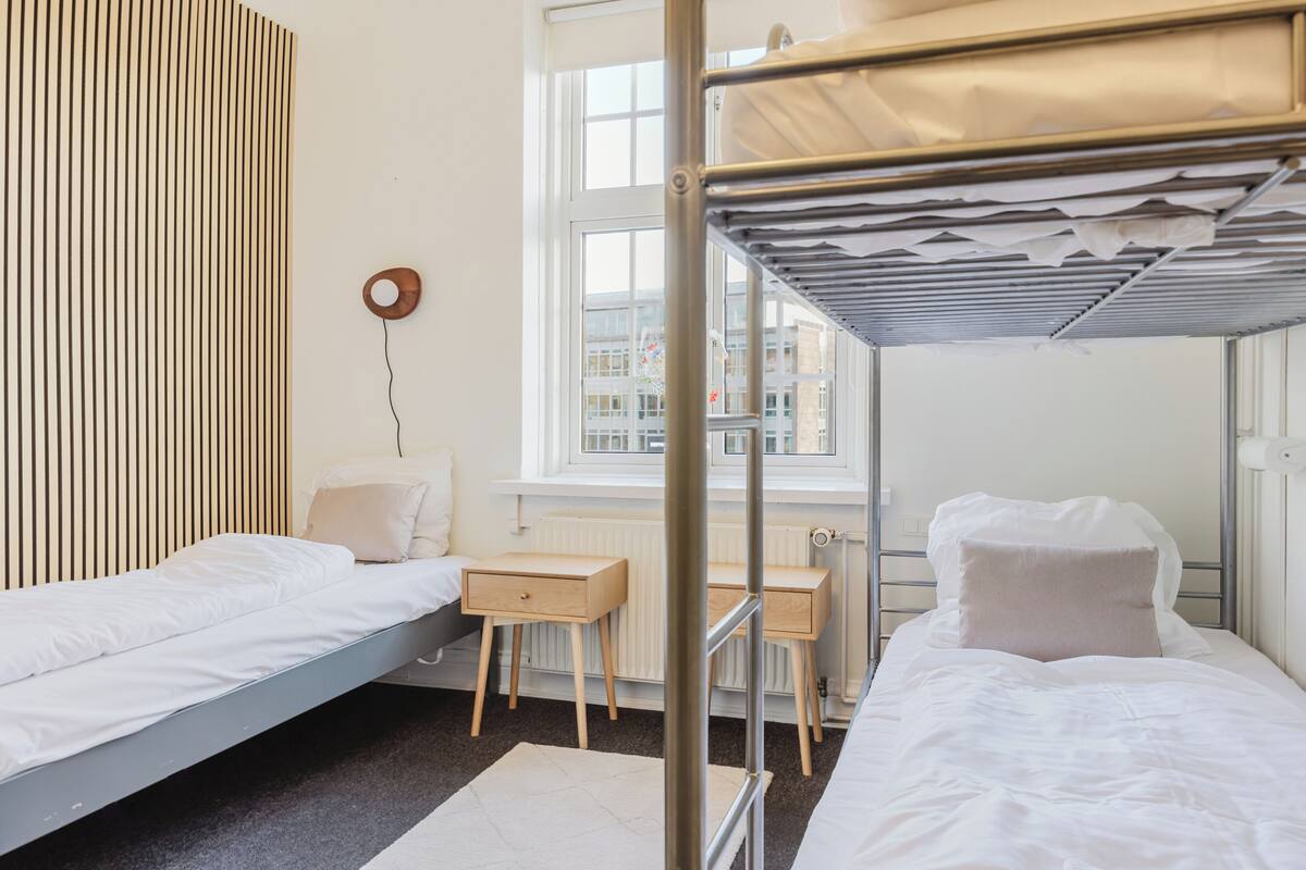 A dormitory room with bunk beds, a desk, a chair, and a radiator.