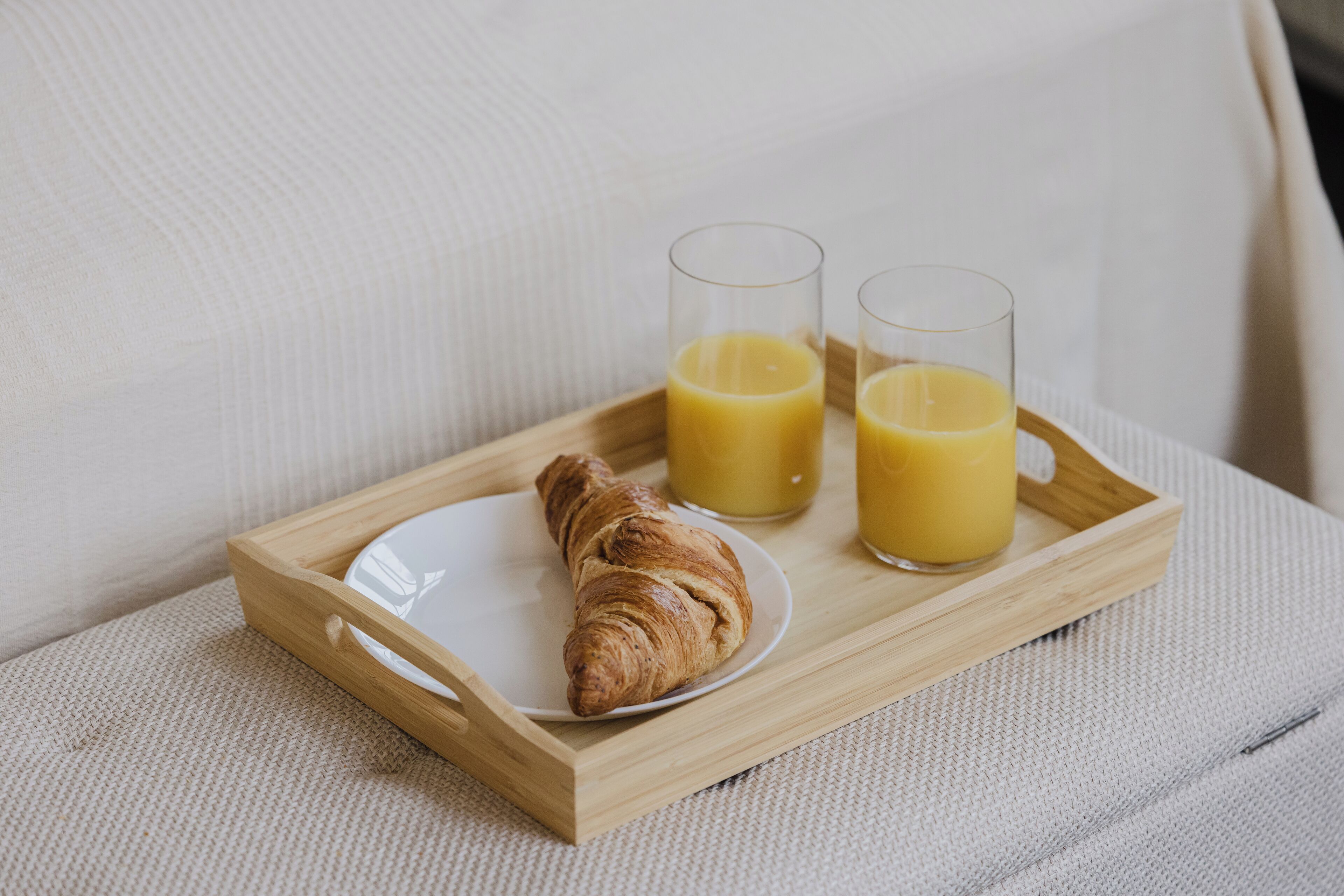 Una bandeja de madera con un croissant y dos vasos de jugo de naranja sobre una superficie blanca con textura.