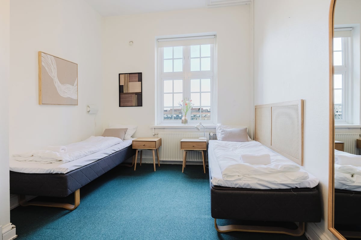 A hotel room with two single beds, a desk, a chair, a window with blinds, and a mirror.