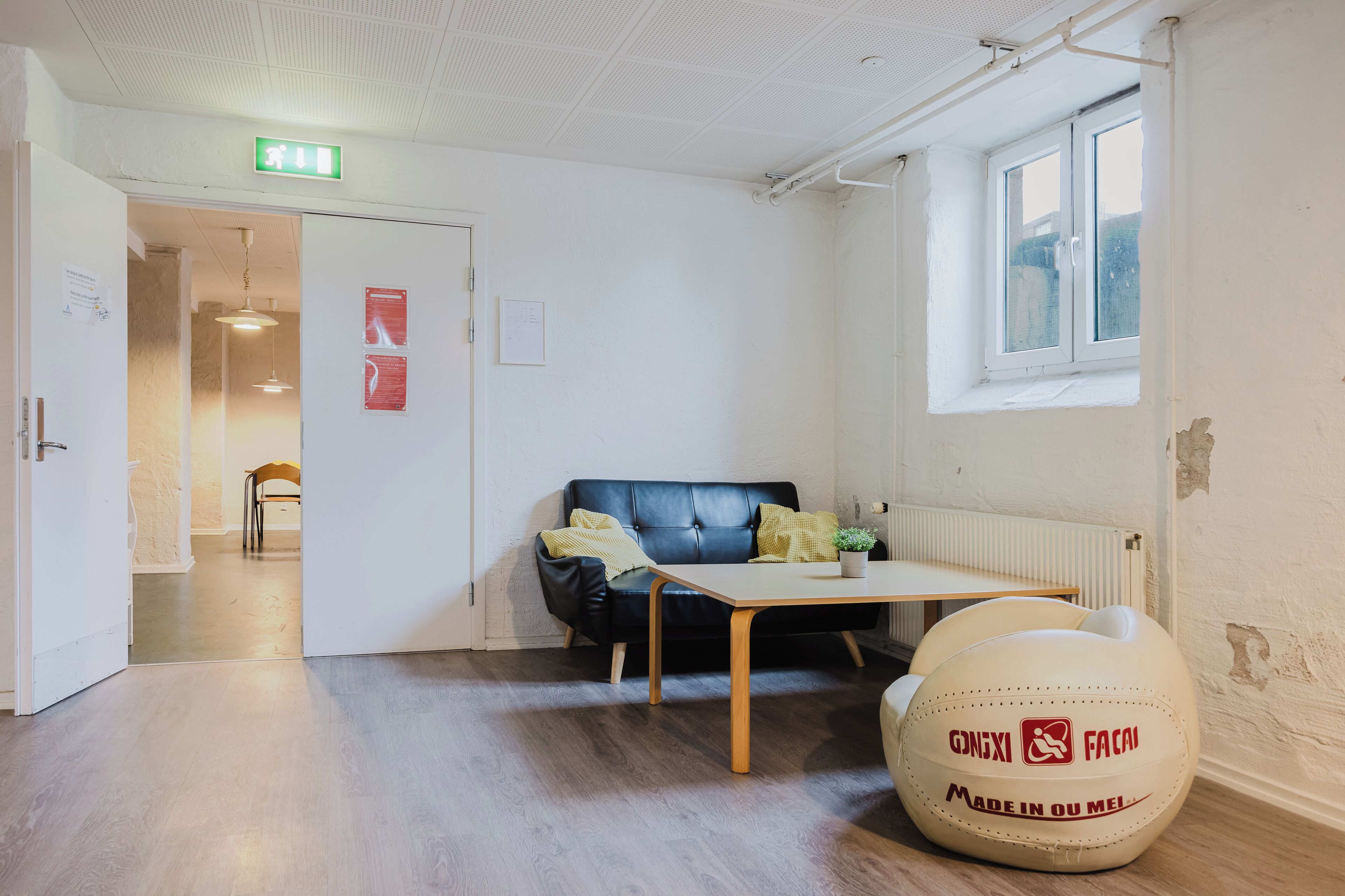 A room with a black leather sofa, a wooden table, and a white bean bag chair. There is a window with a view of the outside, and a door leading to another room.