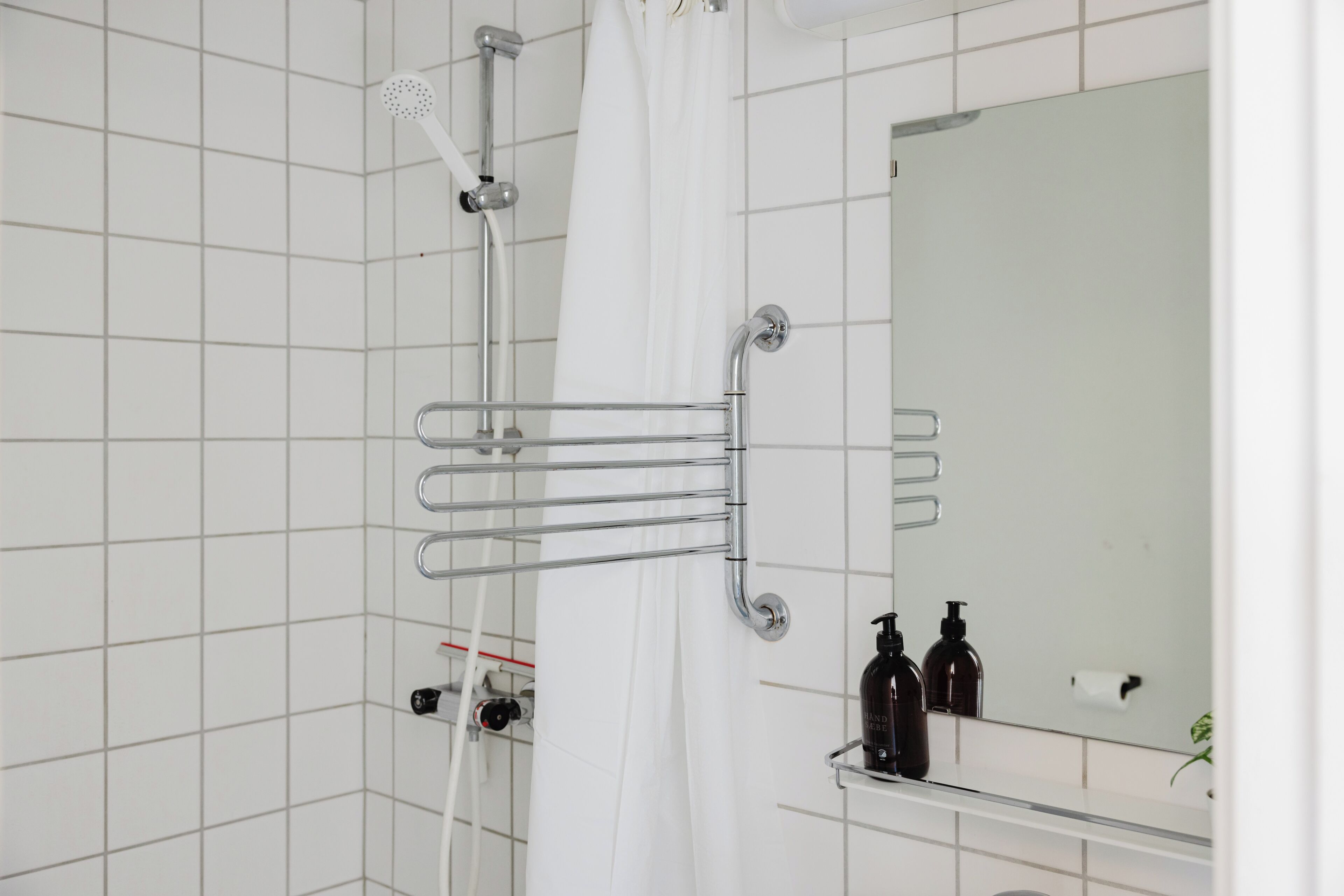 A bathroom with white tiled walls, a shower with a white curtain, a handrail, and a mirror with a shelf holding toiletries.