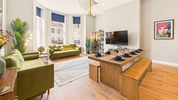 Un salon moderne avec un canapé vert, une table à manger en bois pour six personnes et un grand téléviseur à écran plat fixé au mur.
