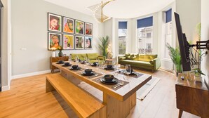 Un espace repas moderne avec une longue table en bois dressée pour un repas, un canapé vert et une grande fenêtre donnant sur des immeubles.