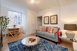 A living room with a grey sofa, a coffee table, a blue patterned rug, a wooden dining table with chairs, and a window with a view of a brick building. - Stylish 2 bed - Tower Hamlets (London)