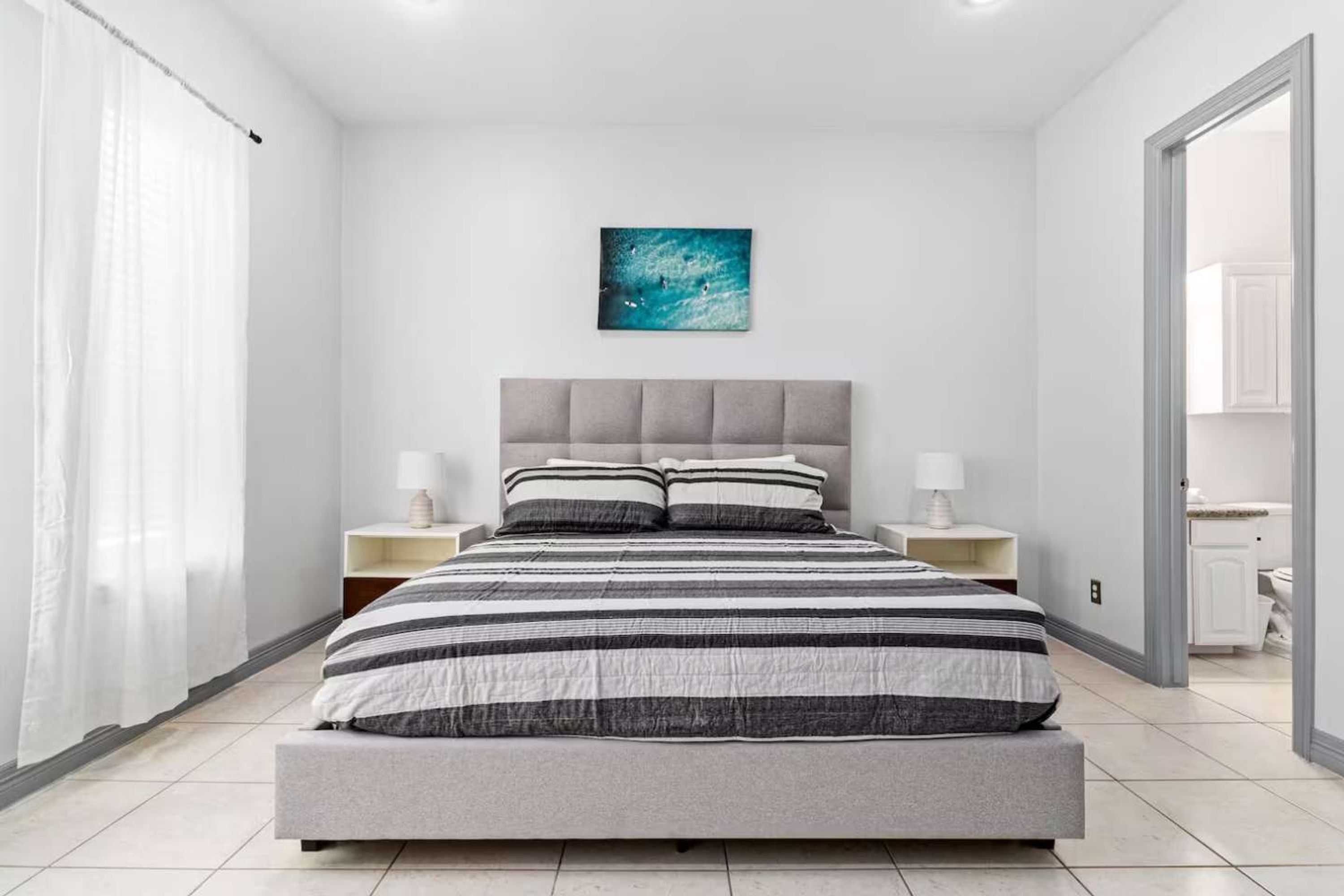 A modern bedroom with a large bed, two bedside tables with lamps, a grey upholstered headboard, a blue artwork on the wall, and a bathroom visible through an open door.